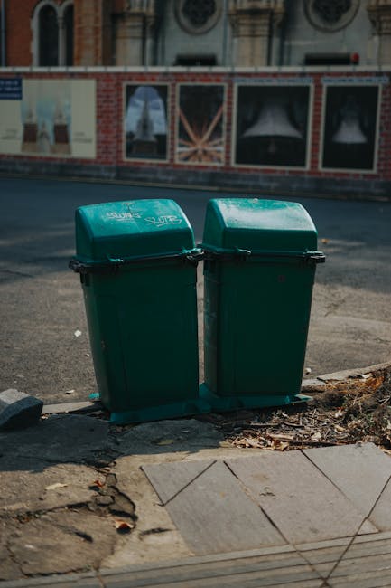 Two dark green, plastic wheelie bins with hinged lids are positioned side by side on a paved sidewalk at the edge of a street, with one slightly in front of the other. The bins are rectangular with rounded edges and a textured surface, featuring black clamp closures across the middle of the lids. They are located near the curb on a section of paving that shows some unevenness and scattered dried leaves and small debris. Behind the bins, the asphalt road appears smooth and slightly reflective under natural daylight, with a faint shadow cast by the bins indicating a light source from the upper left. In the background, there is a brick and stone building with decorative architecture and large windows, suggesting an urban environment. The scene exemplifies a typical setting for external waste receptacles used in private or council rubbish collection services, illustrating a scene where waste clearance or collection is performed away from the property itself, aligned with alternative waste handling methods employed by waste management companies like Waste Clearance Clapham.