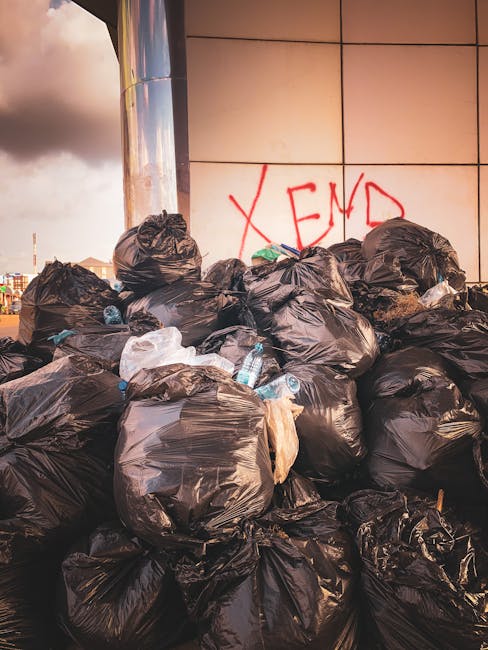 A large pile of black plastic rubbish bags filled with waste is stacked against a tiled wall exterior of a modern building, with some transparent plastic bottles and miscellaneous litter visible among the bags. The bags are tightly knotted at the top and vary in size, occupying most of the foreground and partially obstructing the view of the wall behind. The wall features a prominent red spray-painted graffiti of the word 'XEND' in large, capital letters. The scene is set outdoors, with evening or late afternoon natural lighting casting shadows and a subtle warm hue on the scene. The environment suggests a private or commercial rubbish collection site, perhaps part of an independent waste clearance operation, highlighting the need for professional rubbish removal services. Waste Clearance Clapham's presence in the vicinity indicates efforts to manage or remove this accumulation of waste as part of local rubbish disposal or alternative waste handling solutions.