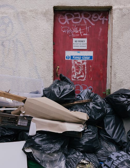 A pile of black rubbish bags and discarded packaging materials stacked against a weathered, beige exterior wall near a red metal door covered in graffiti and stickers, including a white sign indicating 'No Money' and a blue 'Keep Clear' notice, suggesting a scene of waste accumulation outside a building. The rubbish bags are made of plastic and are loosely tied, with some items such as cardboard and plastic sheets visibly protruding, indicating collection for waste removal services. The surrounding environment appears to be an urban alley or side street, with a concrete ledge and minimal greenery. The scene exemplifies typical waste awaiting private rubbish clearance, highlighting the importance of professional waste management, like the services offered by Waste Clearance Clapham, for efficient and responsible disposal in residential or commercial settings. The lighting is natural and diffuse, typical of overcast conditions, providing a neutral, clear view of the cluttered scene without shadows or glare.