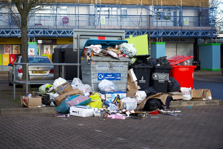 A collection of overflowing rubbish bins and scattered waste bags is situated on a paved sidewalk in front of a commercial building with blue scaffolding and a fish bar. The waste includes a large metal recycling bin labeled for mixed paper and cardboard, which is open and filled with crumpled paper and cardboard boxes. Surrounding the bin are black and red plastic waste bags, some torn open, with various discarded packaging, papers, and cardboard boxes spilling onto the ground. A small grey car is parked nearby, partly obscured by a metal railing separating the waste area from the vehicle. In the background, the building has storefronts with bright signage and yellow accents, and the scene is lit by natural daylight. The mess suggests a situation of unscheduled or improper waste disposal, which might be addressed through private rubbish removal services like those offered by Waste Clearance Clapham, supporting alternative on-site waste management solutions.