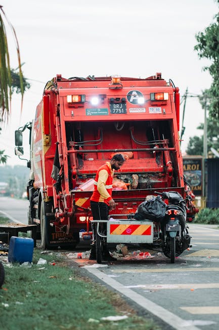 A waste management worker in a yellow and red uniform is seen standing on a street, collecting debris from the rear section of a large red rubbish collection truck. The truck, branded with a company logo and identification number, is positioned on the roadside amid a suburban or urban environment, with trees and power lines visible in the background. The worker appears to be handling black plastic bags placed near the truck's rear hopper, which is open and ready for waste intake. The scene shows some scattered litter on the ground, along with a small wheelie bin nearby, indicating ongoing rubbish collection or clean-up activity. The lighting suggests daytime with overcast sky, and the surroundings appear to be a residential or commercial area, consistent with private waste handling services independent of local authority collection, as often seen in alternative rubbish removal or on-site clearance situations. Waste Clearance Clapham is identifiable as the service provider responsible for this operation.