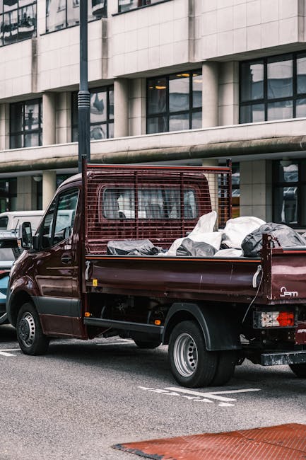 A dark red flatbed truck parked on a city street in front of a modern multi-story building with large glass windows and a concrete facade. The truck's cargo area is filled with various types of waste materials, including black and white bags, some of which appear to be filled with household or construction debris. The truck has a metal safety grille at the front of the cargo bed and is positioned close to the curb, with a section of street markings visible in the foreground. A white vehicle is partially visible parked nearby. The scene, captured during daylight, suggests an on-site or private waste collection process typical of rubbish removal services, such as those offered by Waste Clearance Clapham, and reflects the practical aspect of alternative waste handling outside local authority disposal methods.